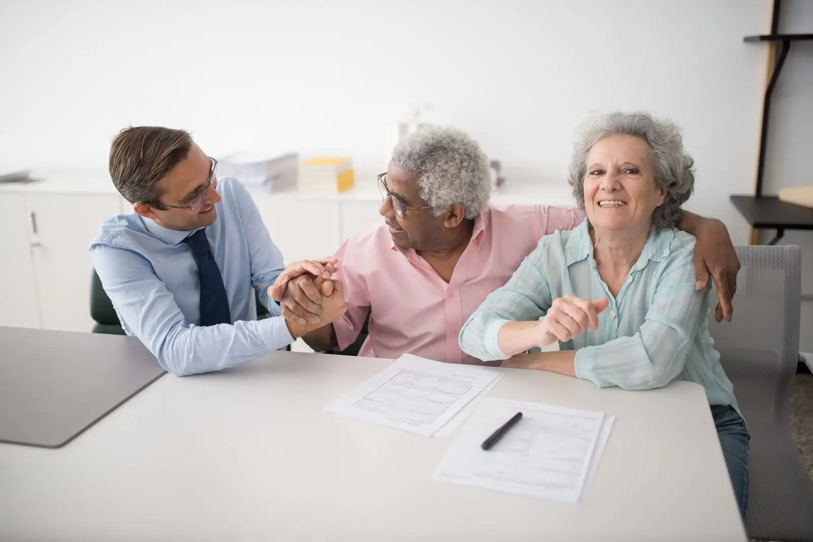 Conseiller financier serrant la main d'un couple de seniors souriants autour de documents d'épargne sur un bureau