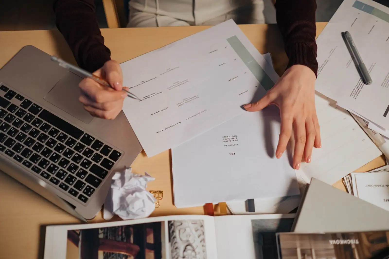Personne travaillant sur un ordinateur portable, stylo en main, entourée de documents papier sur un bureau