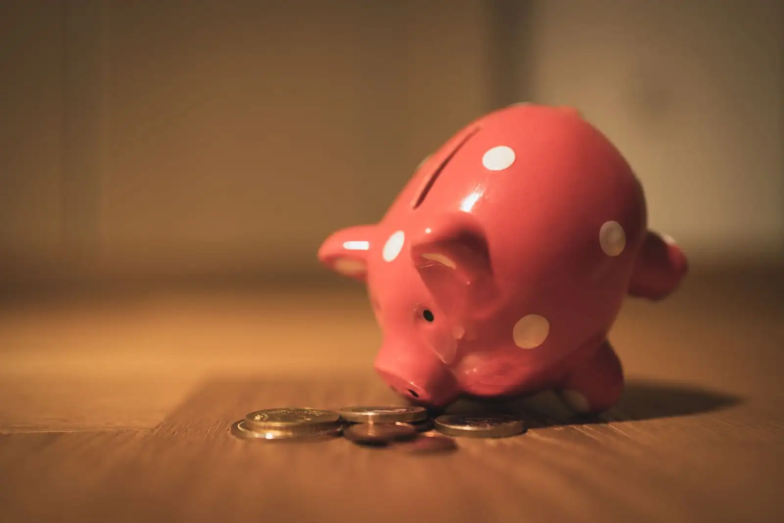 Tirelire en céramique rose à pois blancs posée à côté de quelques pièces de monnaie sur un sol en bois