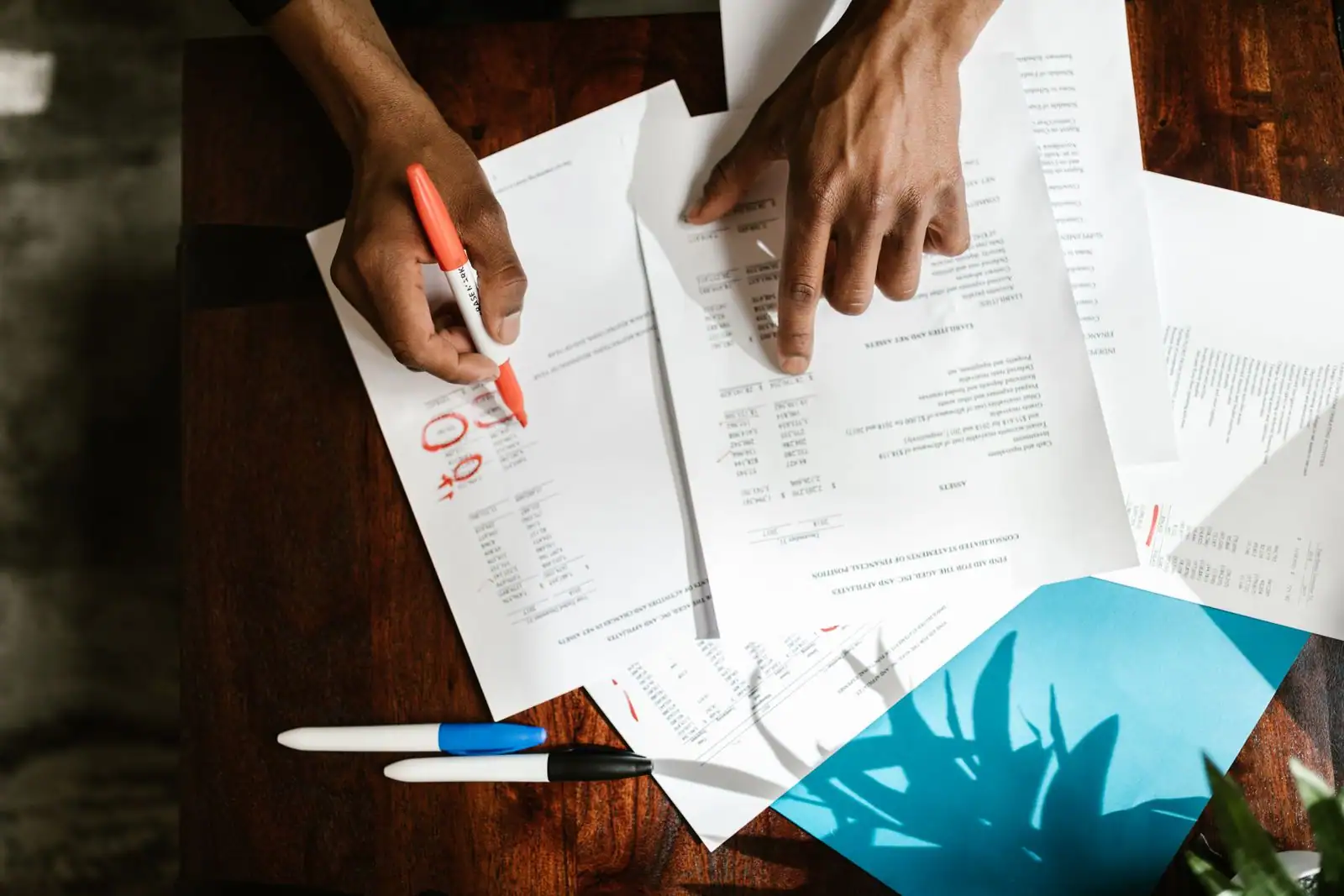 Des mains annotent au feutre des documents financiers chiffrés posés sur un bureau en bois