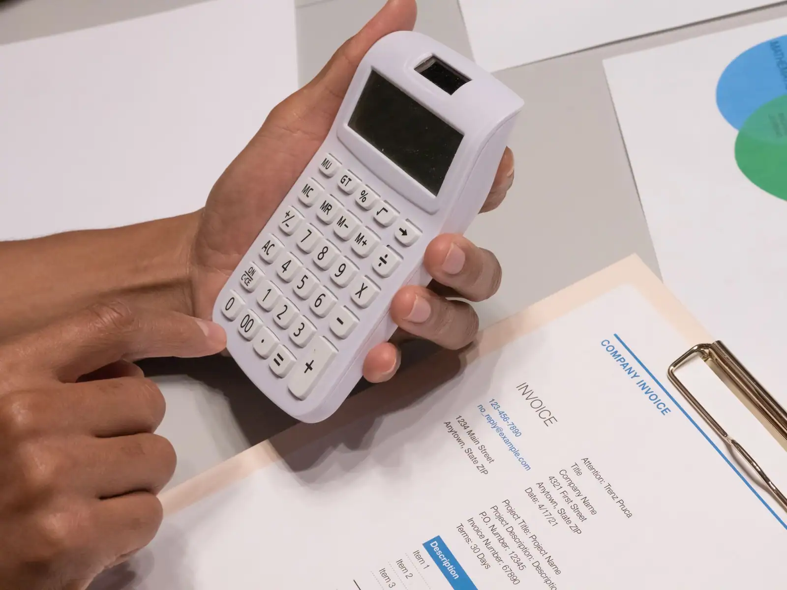 Personne vérifiant une facture d'entreprise à la calculatrice, documents et graphiques posés sur le bureau.