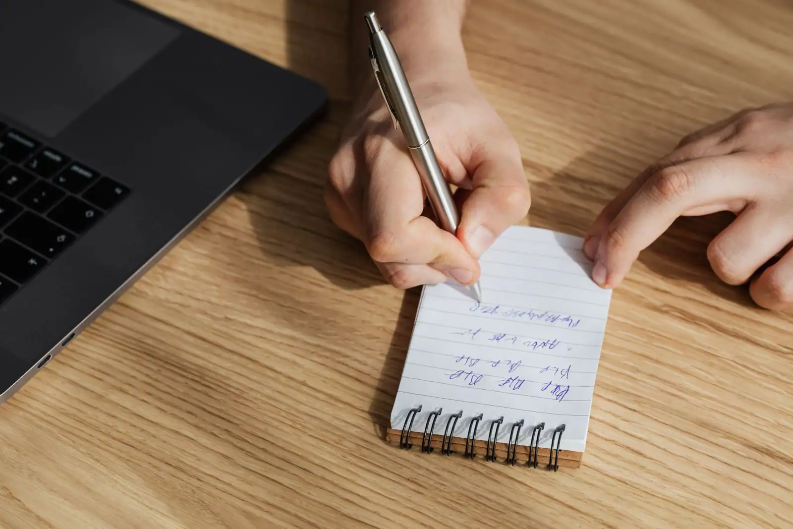 Une main écrit des notes sur un carnet à spirale posé près d'un ordinateur portable, sur un bureau en bois.