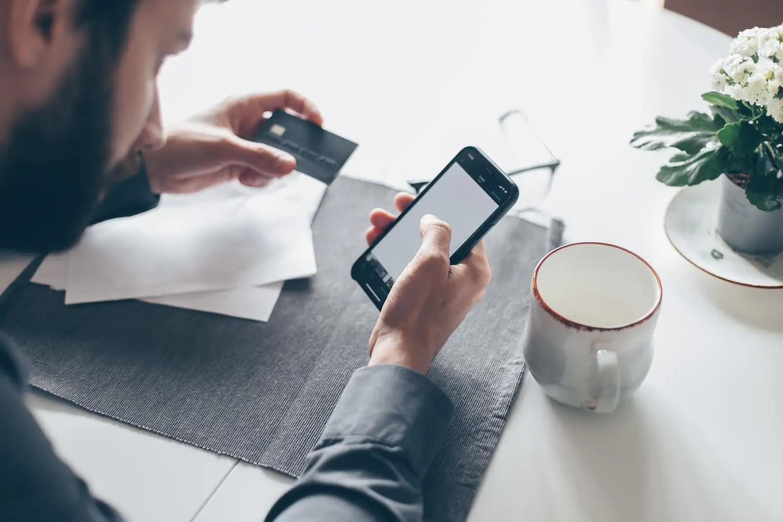 Personne attablée chez elle, carte bancaire dans une main et smartphone dans l’autre, papiers posés à côté