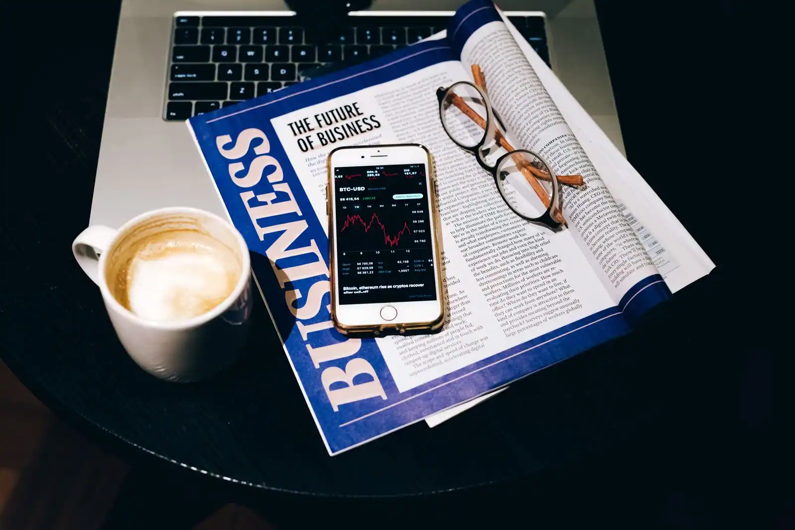 Un magazine économique en anglais ouvert et un smartphone affichant un graphique de marché, à côté d'un café.